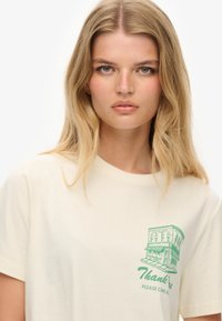 Cremefarvet bomulds T-shirt med en grøn grafisk illustration af en butik og teksten "Tak, KOM VENLIGST AL." Enkel, afslappet pasform.