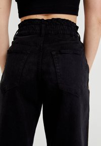 Svarta denimbyxor med volang i midjan, har två bakfickor och rak benlinje. Matt ytstruktur och minimalistisk stil.