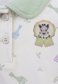 Polo in cotone bianco con colletto verde, con motivi di giraffe pastello. Include una toppa con una giraffa sorridente e il testo "Sophie la girafe".