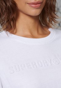 Witte katoenen t-shirt met een ronde halslijn, voorzien van reliëf "SUPERDRY" branding aan de voorkant, een gladde textuur en korte mouwen.