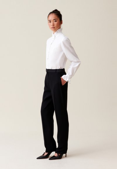 Blusa bianca con colletto alto a volant e chiusura frontale con bottoni, abbinata a pantaloni neri sartoriali e scarpe nere con tacco a punta decorate con fiocchi.