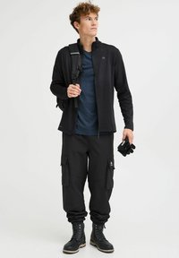 Giacca nera con zip sopra una t-shirt blu navy, abbinata a pantaloni cargo neri con tasche. Stivali neri con lacci e guanti.