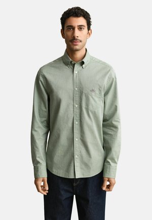 Man met kort krullend haar, gekleed in een lichtgroen overhemd met knopen en donkere spijkerbroek, staand tegen een effen achtergrond.