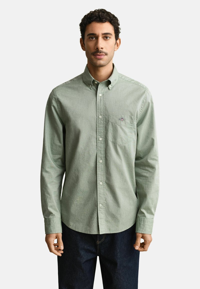 Hombre con cabello corto y rizado, vistiendo una camisa de botones verde claro y jeans oscuros, de pie frente a un fondo liso.