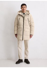 Beige pufferjack met kap, voorzien van een ritssluiting aan de voorkant, twee grote zakken en zwarte accenten. Gecombineerd met crèmekleurige broek en donkere schoenen.