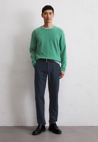 Grüner Strickpullover mit weißem Kragen, kombiniert mit dunklen grauen Hosen und schwarzen Schnürschuhen. Das Outfit hat eine lässige Passform.