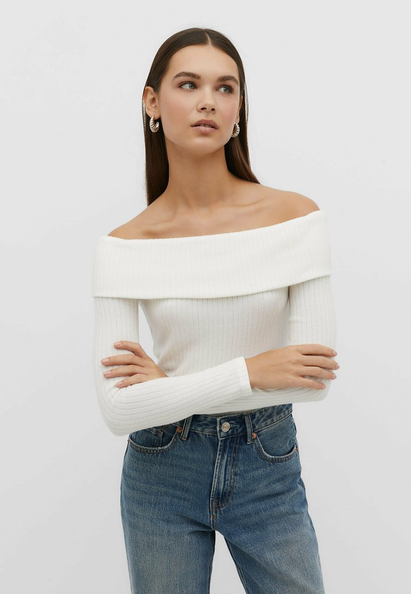 Off-Shoulder weißes Ripp-Top mit langen Ärmeln, kombiniert mit hochtaillierten blauen Jeans aus Denim, die einen lässigen Schnitt und eine subtile Textur aufweisen.