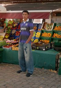 Ung mand i en fodboldtrøje og slidte jeans står med hænderne i lommerne ved farverige frugttrækasse ved en udendørs markedstand.