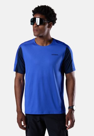 Uomo che indossa occhiali da sole sportivi riflettenti, maglietta a maniche corte blu Rossignol con pannelli laterali scuri, pantaloni neri e orologio da polso nero.