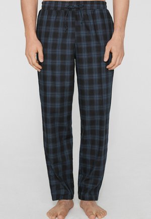 Hombre con pantalones de pijama de cuadros negros y azules, de pie descalzo frente a un fondo claro y liso.