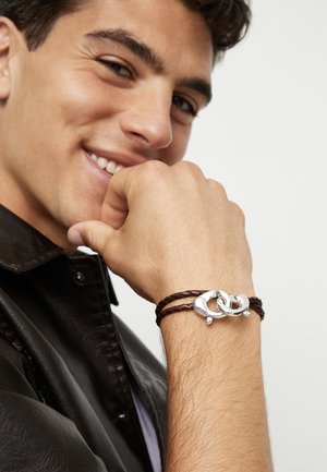 Pulsera trenzada de cuero marrón con eslabones plateados entrelazados y un cierre elegante. Se lleva en una mano con una chaqueta de cuero.