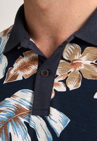 Polo de manga corta con base azul marino y grandes patrones florales multicolores. Incluye un cuello oscuro y un detalle de botón.