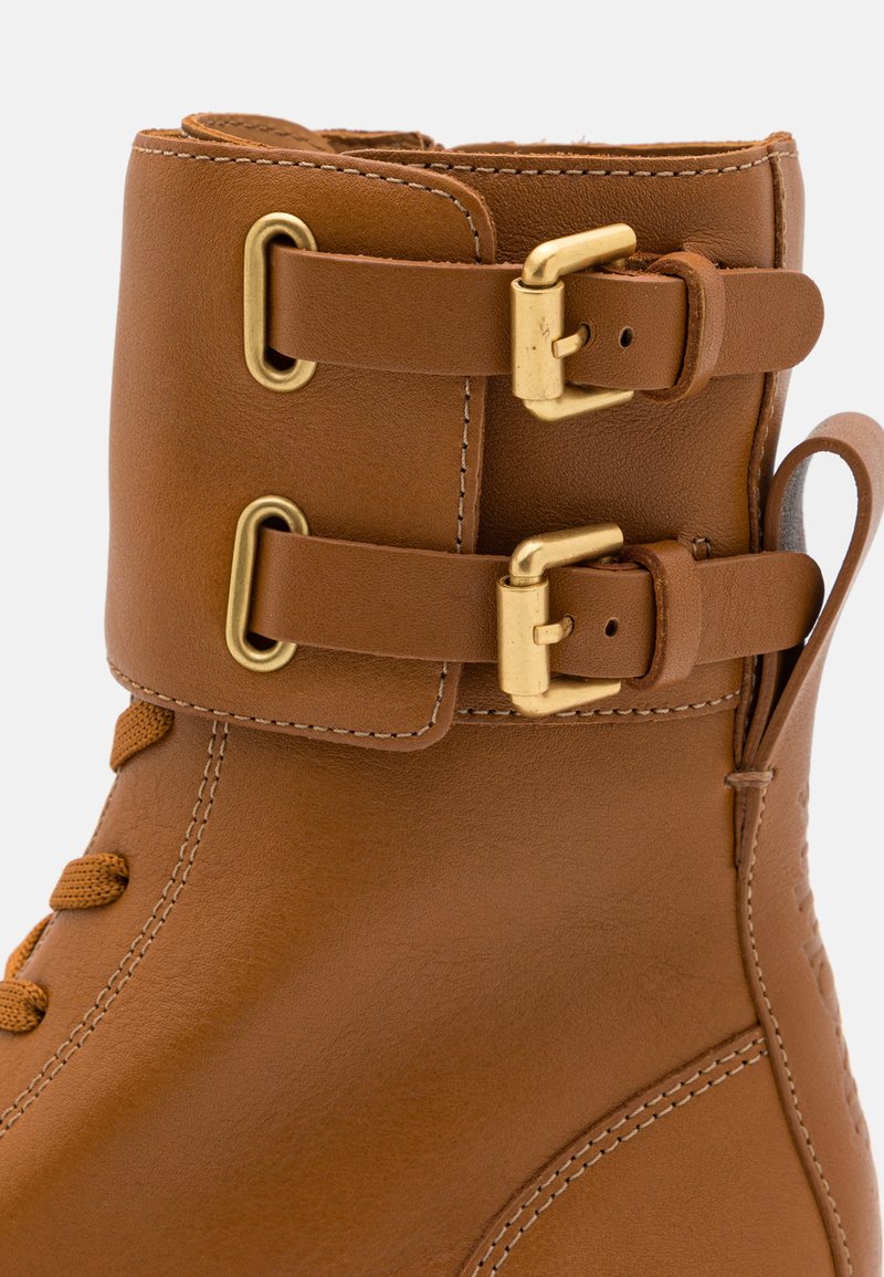 Stivale alla caviglia in pelle marrone con doppie fibbie dorate, superficie testurizzata e cuciture dettagliate. Punta rotonda e chiusura a lacci sicura.