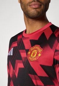 Rot und schwarz gestreiftes Kurzarmtrikot mit Zickzackmuster. Mit Rundhalsausschnitt und dem Manchester United-Logo in Gelb und Rot auf der Brust.
