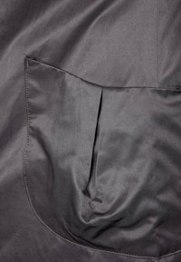 Il tessuto grigio con una texture liscia presenta una grande tasca curva in basso a sinistra. La tasca ha sottili linee orizzontali nel materiale.