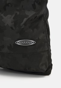 Tissu noir à motif camouflage avec coutures surpiquées et logo ovale blanc indiquant "URTRN" près du bord inférieur.