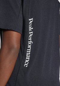 Sort kortærmet t-shirt lavet af blødt stof, med hvid tekst "Peak Performance" langs sidesømmen. Glat tekstur, afslappet design.
