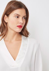 Blusa blanca con un escote en V profundo y ribete de satén. Tejido suave con un corte relajado y mangas largas. Diseño simple y elegante.