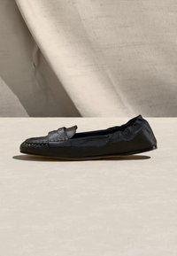 Mocassin plat en cuir noir avec une tige texturée et un dos élastiqué, présenté sur une surface beige clair avec un fond en tissu.