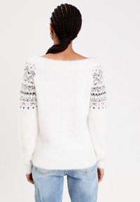 Personne avec des cheveux tressés porte un pull en laine blanc avec des manches à motifs et un jean bleu clair, vu de dos sur un fond blanc.