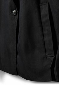 Gros plan d'une veste noire montrant un bouton gris sur le devant et une poche fendue verticale sur le côté.