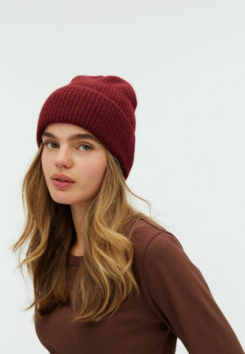 Căciulă tricotată burgundy cu o textură moale, având un model rilbat. Designul este lejer, permițând o potrivire confortabilă.