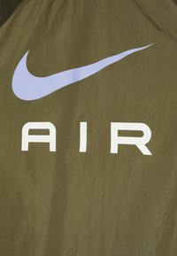 Olivově zelená bunda s texturovaným povrchem, na které je velký bílý nápis "AIR" a světle fialové logo Nike swoosh.