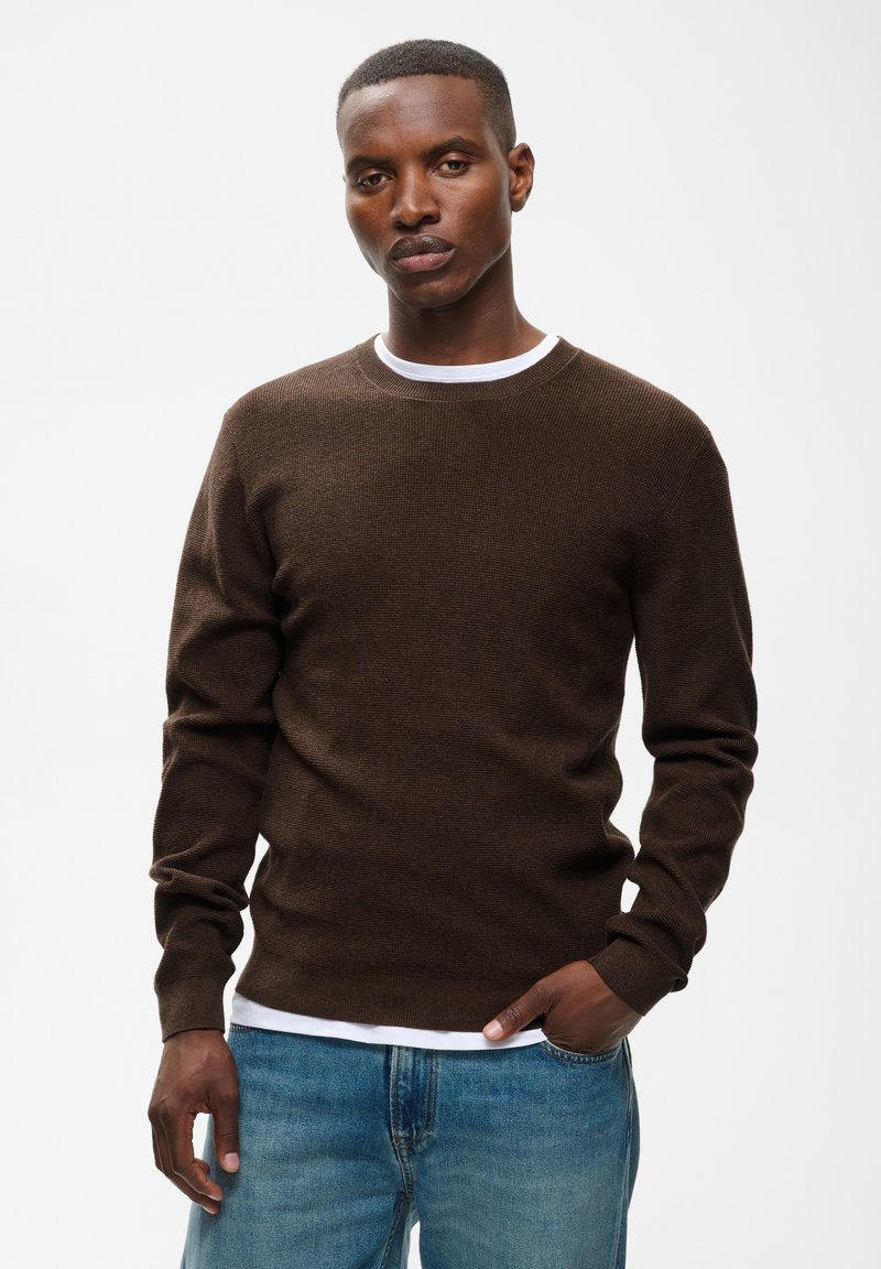 Homme portant un pull marron foncé sur une chemise blanche et un jean bleu, debout avec une main dans la poche devant un fond blanc uni.