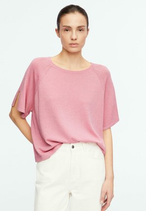 Frau mit zurückgebundenen dunklen Haaren, die ein locker sitzendes rosa Kurzarmoberteil und hoch taillierte weiße Hosen vor einfarbigem Hintergrund trägt.