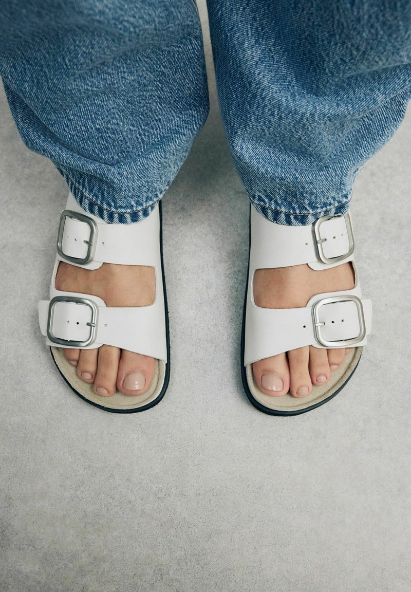 Sandali bianchi con due fibbie argentate, design open toe, suola nera a contrasto, indossati con jeans in denim blu. Stile piatto e casual.