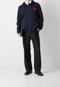 Hombre con sudadera azul marino sobre camisa blanca y corbata negra, pantalones negros, sosteniendo un clutch negro, de pie sobre un fondo blanco liso.