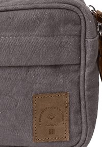 Sac bandoulière en tissu gris de forme rectangulaire, avec une étiquette en cuir marron et une surface texturée. Comprend une poche avant.