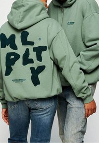 Des sweat-shirts verts avec un grand logo "M L P L T Y" en peluche au dos et un texte plus petit en dessous. Associés à un jean bleu, affichant un style décontracté.