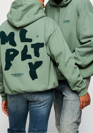 Des sweat-shirts verts avec un grand logo "M L P L T Y" en peluche au dos et un texte plus petit en dessous. Associés à un jean bleu, affichant un style décontracté.