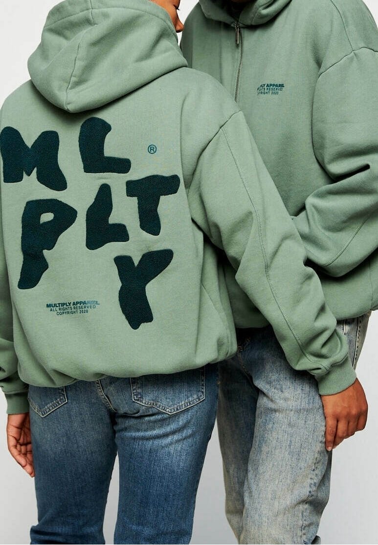 Des sweat-shirts verts avec un grand logo "M L P L T Y" en peluche au dos et un texte plus petit en dessous. Associés à un jean bleu, affichant un style décontracté.