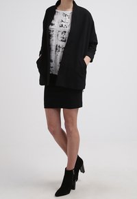 Svart kappa med öppnatch fram, med två fickor, i kombination med en mönstrad vit och svart topp samt en svart kjol, kompletterad med svarta ankelskor.