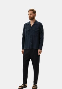 Camisa de botones azul marino con cuello, dos bolsillos en el pecho y un corte relajado, combinada con pantalones negros ajustados y sandalias negras.