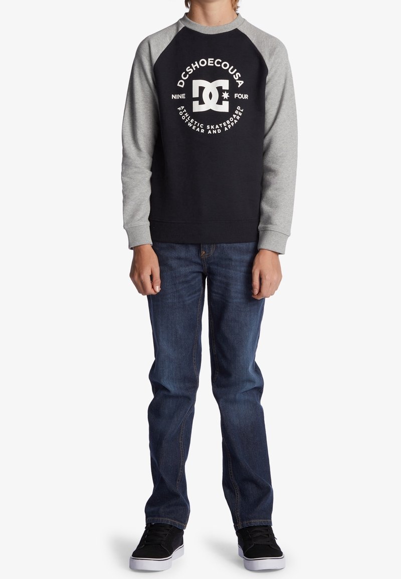 Sweatshirt bleu marine avec manches raglan grises, arborant un logo blanc et un motif texte. Porté avec un jean bleu et des baskets noires.