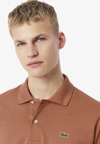 Brun polo-skjorta med en strukturerad tygkvalitet, som har en tvåknapps knäppning och en liten grön krokodil-logotyp på vänster bröst.