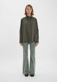 Chemise oversized à carreaux verts avec un devant à boutons, associée à un pantalon évasé vert clair et des chaussures noires, sur un fond neutre.