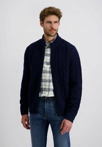 Marineblauer Zopfmuster-Cardigan mit Stehkragen und gerippten Bündchen. Getragen über einem karotten Hemd und kombiniert mit blauen Jeans.