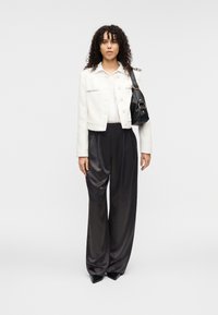 Λευκό cropped σακάκι με υφή και ασημί λεπτομέρειες, σε συνδυασμό με μαύρο σατινέ παντελόνι με φαρδιά μπατζάκια και μυτερά μαύρα παπούτσια, κρατώντας μια μαύρη τσάντα χειρός.