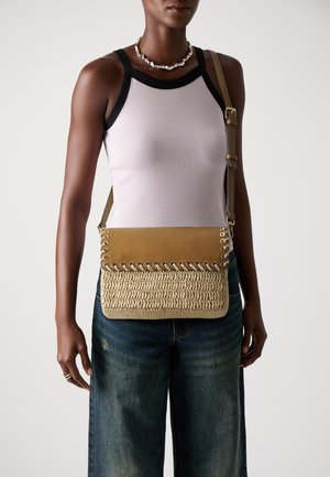Frau in lila ärmellosem Oberteil und Jeans, die eine beige geflochtene Umhängetasche mit Lederklappe und -riemen trägt.