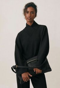 Czarna, oversize bluza z wysokim kołnierzem i dekoltem w kształcie litery V, w zestawie z czarną skórzaną torbą na ramię z zamkiem błyskawicznym.