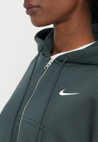 Zeleni dukserica sa zatvaračem, izrađena od mekanog materijala, s bijelim Nike logom na prsima i kapuljačom sa vezicama. Prednja kenguru džep.