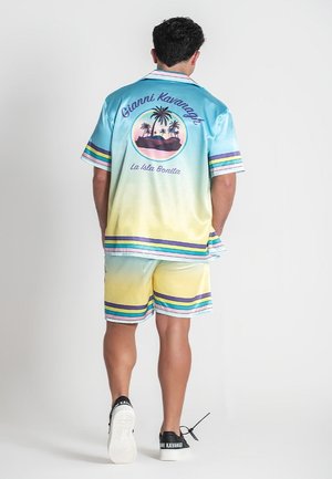 Mann trägt ein blau-gelbes Ombre-Kurzarmhemd und passende Shorts mit Palmenmotiv und dem Text "Gianni Kavanagh La Isla Bonita" auf der Rückseite.
