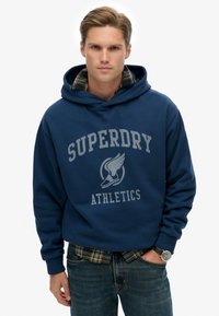 Marineblauwe hoodie met grijze "Superdry Athletics" tekst, kangaroozak en ribgebreide manchetten, over een geruite shirt gelaagd. Spijkerbroek, polshorloge zichtbaar.