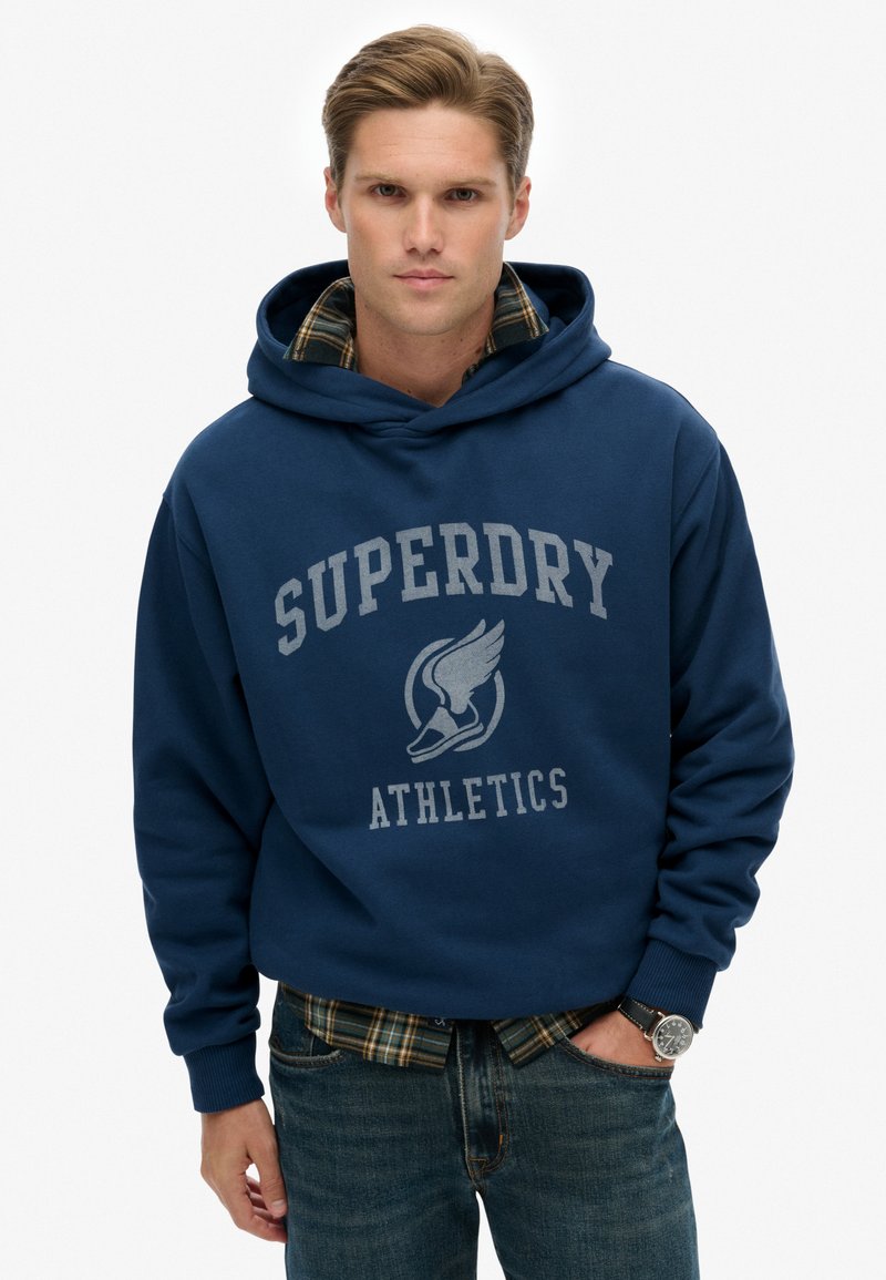 Marineblauwe hoodie met grijze "Superdry Athletics" tekst, kangaroozak en ribgebreide manchetten, over een geruite shirt gelaagd. Spijkerbroek, polshorloge zichtbaar.
