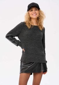 Dunkelgrauer gestrickter Pullover mit geripptem Saum und lederähnlichen Ärmeln, kombiniert mit einem schwarzen Rock aus Kunstleder. Das Modell trägt eine schwarze Mütze.