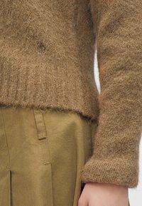Manche et ourlet d'un pull en maille marron duveteuse sur un pantalon vert olive avec passants de ceinture, montrant un gros plan de l'avant-bras et de la taille d'une personne.
